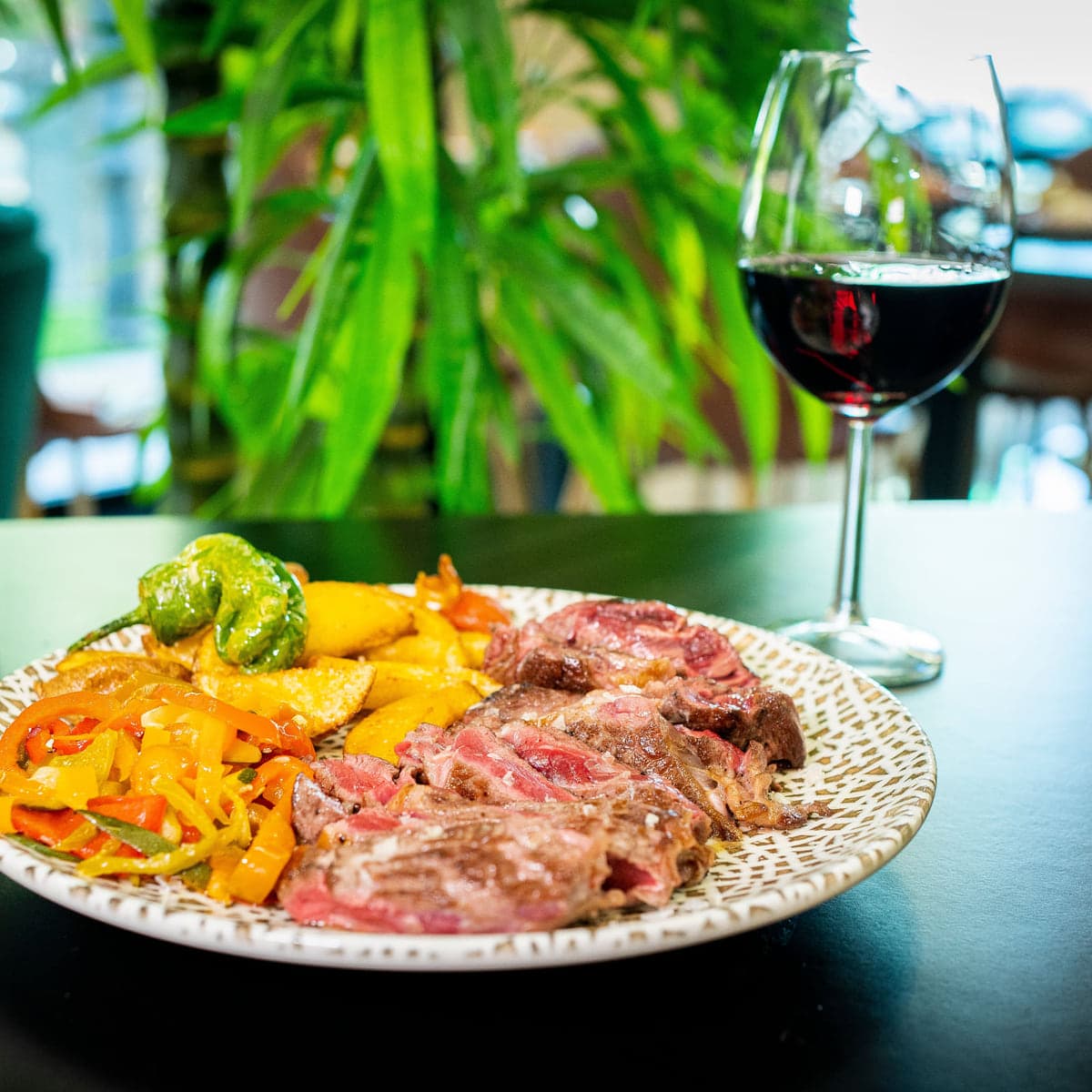 Carne en lonchas con pimientos asados y patatas, junto a una copa de vino tinto