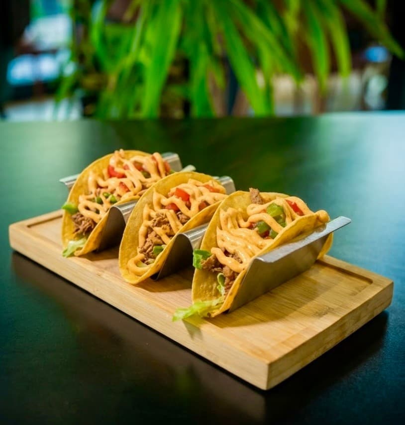 Tres tacos de cochinita pibil en su soporte, sobre una tabla de madera
